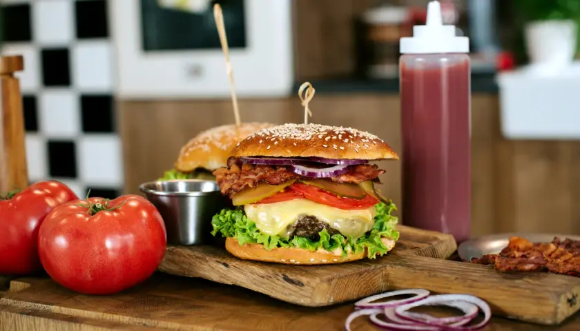 Soczysty burger wołowy z bekonem, serem i warzywami na desce, obok pomidorów i sosu.