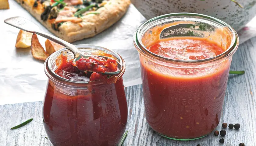 Dwa słoiki domowego sosu do pizzy, jeden otwarty z łyżką pełną aromatycznej masy, obok kawałek pizzy i przyprawy.