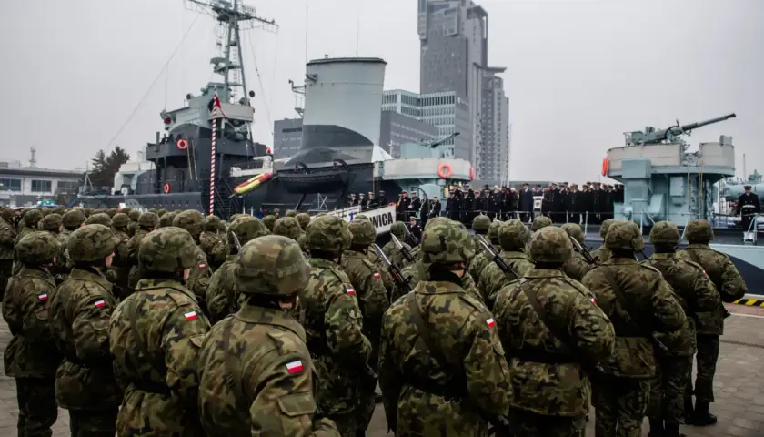 Żołnierze w mundurach polowych stoją w szyku przed okrętem wojennym. W tle nowoczesne budynki i inni wojskowi. To część ćwiczeń wojskowych.