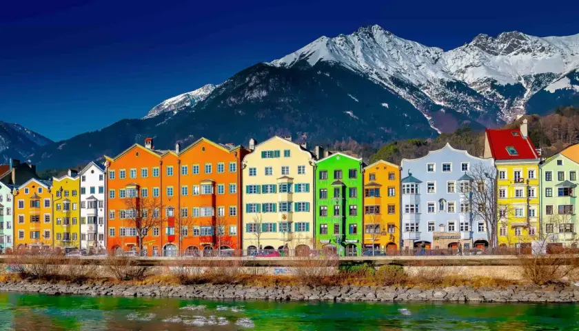 Colorful buildings line the Inn River, a vibrant scene on the journey from St. Moritz to Innsbruck, with snow-capped mountains in the background.