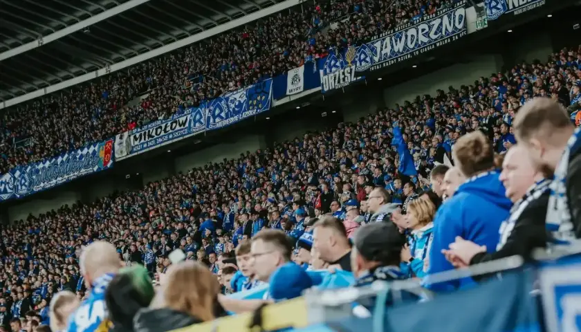 Tłum kibiców na stadionie, w niebieskich barwach, z transparentami "Jaworzno", "Mikołów", "Chorzów". Wszyscy w zgodzie dopingują swoją drużynę.