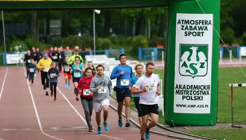Uczestnicy biegną pod zieloną bramą z napisem "Akademickie Mistrzostwa Polski". Wszyscy biorą udział w teście coopera, pokazując ducha sportowej rywalizacji.