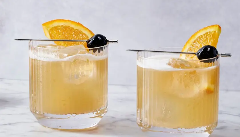 Dwa drinki Rusty Rat z pomarańczą i wiśnią na patyczku, podane w szklankach z lodem.