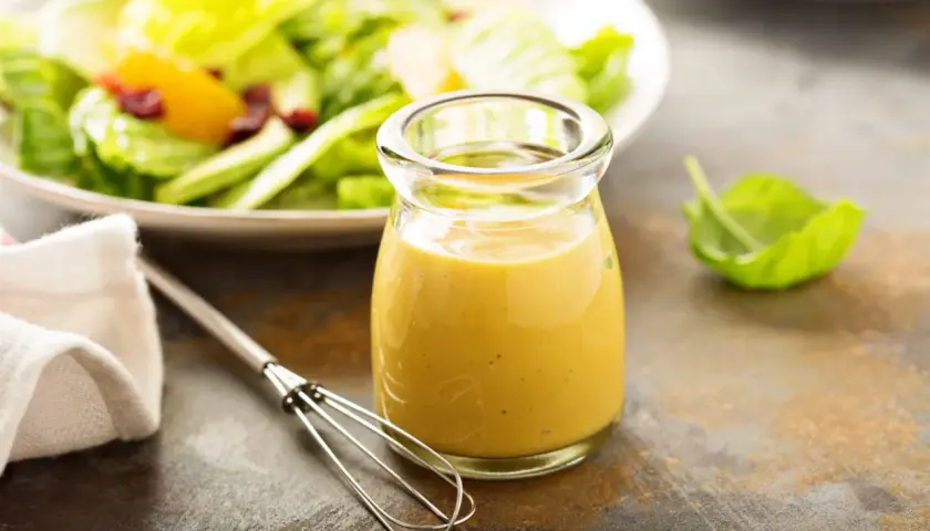 Ein Glas mit gelbem Blattsalat Dressing steht neben einem Schneebesen. Im Hintergrund ein Teller mit frischem Salat.