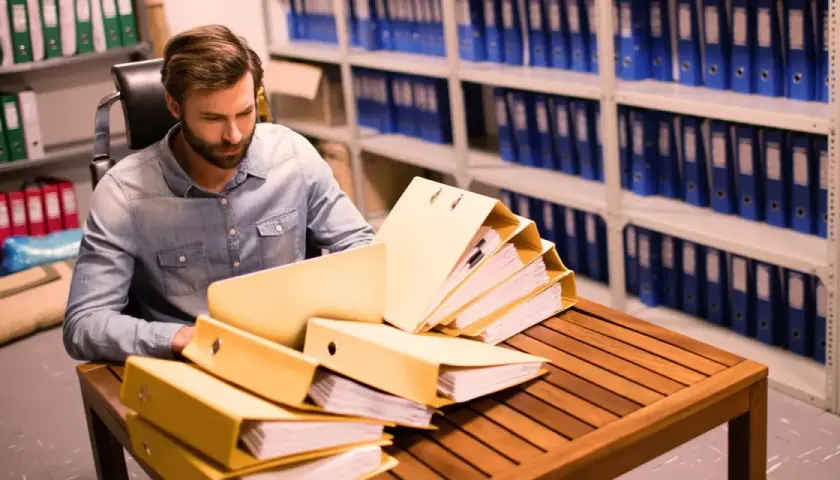 Un homme examine des dossiers dans un **archive papier**. Des étagères remplies de classeurs bleus et verts s'étendent à l'arrière-plan.