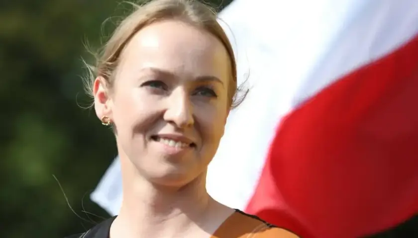 Uśmiechnięta Urszula Brzezińska-Hołownia, której wiek nie jest widoczny, na tle polskiej flagi.