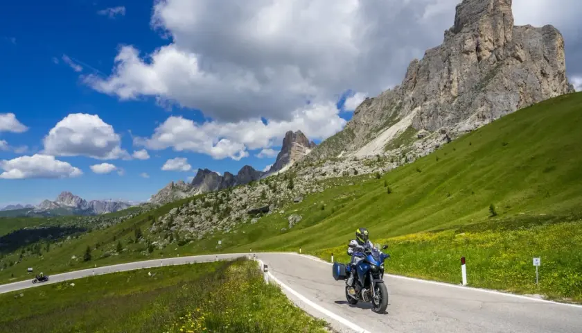 Motociclista percorre la strada cimina tra prati verdi e montagne imponenti sotto un cielo azzurro con nuvole bianche.