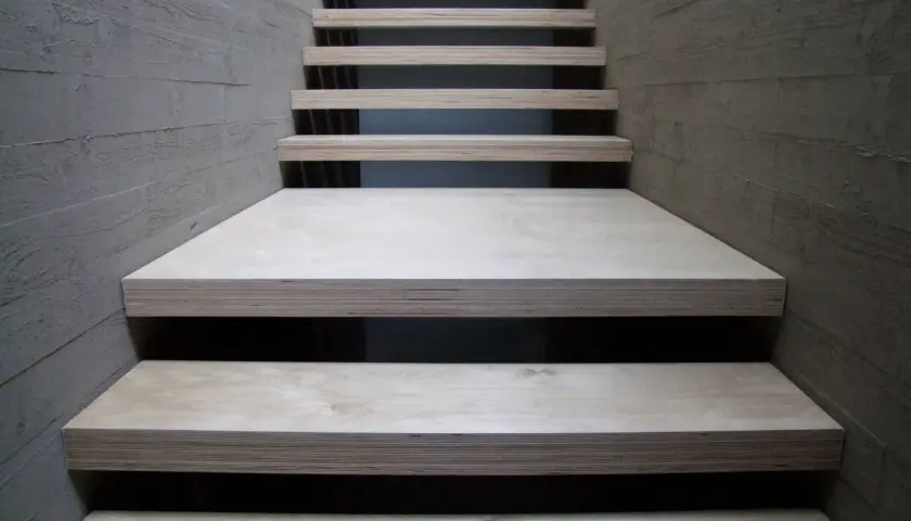 Una scala moderna con gradini in legno compensato, materiale resistente e versatile, fiancheggiata da pareti in cemento grezzo.