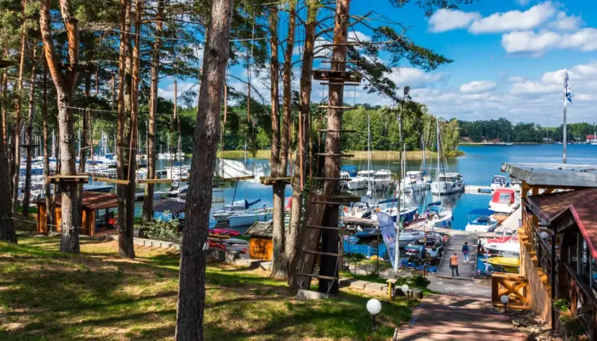 Linowy park rozrywki w lesie nad jeziorem z przystanią pełną jachtów.
