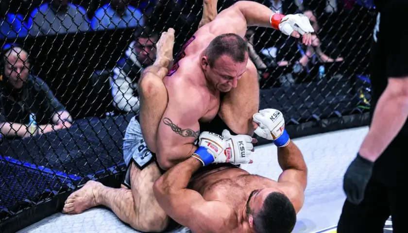 Intensywna walka Pudziana w klatce MMA. Jeden zawodnik klęczy, drugi próbuje go obalić.