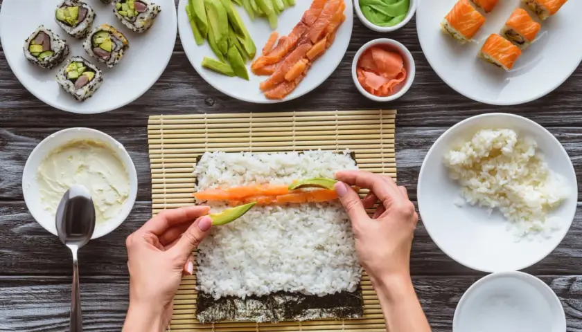 Händer lägger lax och avokado på sushiris för ett sushirecept. Ingredienser som gurka, wasabi och färdig sushi syns också.