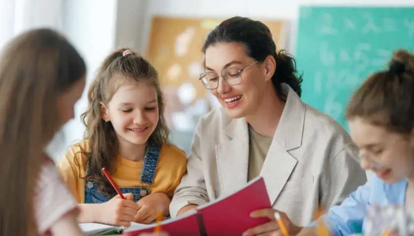 Lehrerin gibt Schülern bei einer Klassenleiterstunde Ideen. Sie schauen gemeinsam in ein Buch, während die Kinder schreiben.