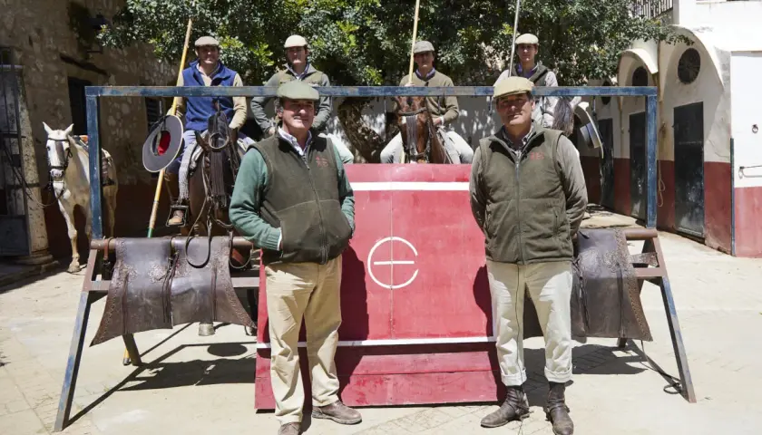 Hombres con gorras y chalecos posan frente a una barrera roja. Uno de ellos, Julio de la Puerta, sonríe. Caballos y equipo de equitación completan la escena.
