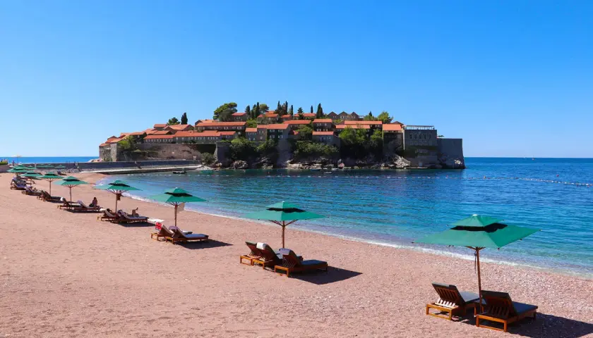 Sonnige Bucht mit rotem Sandstrand und Liegen am Meer in Montenegro. Im Hintergrund die historische Insel Sveti Stefan.