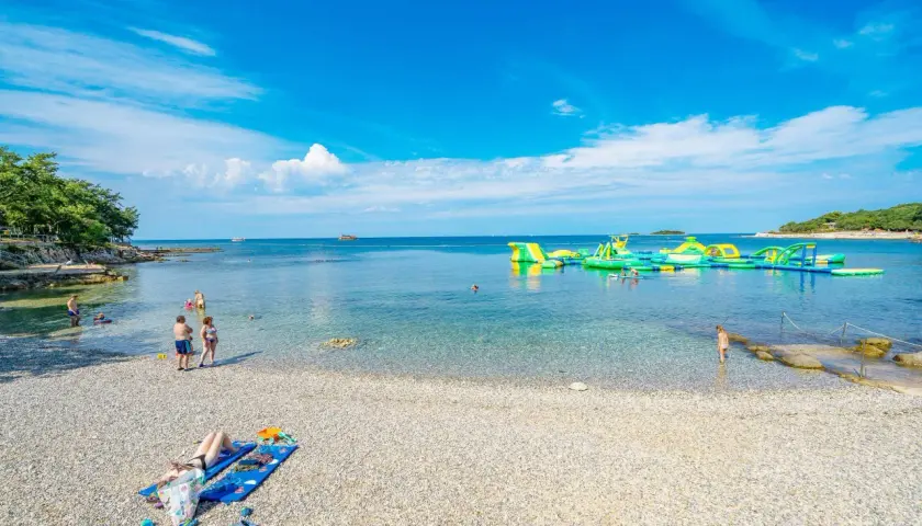 Słoneczna plaża Bijela Uvala z kamyczkami, turkusową wodą i dmuchanymi atrakcjami wodnymi. Ludzie odpoczywają i kąpią się.