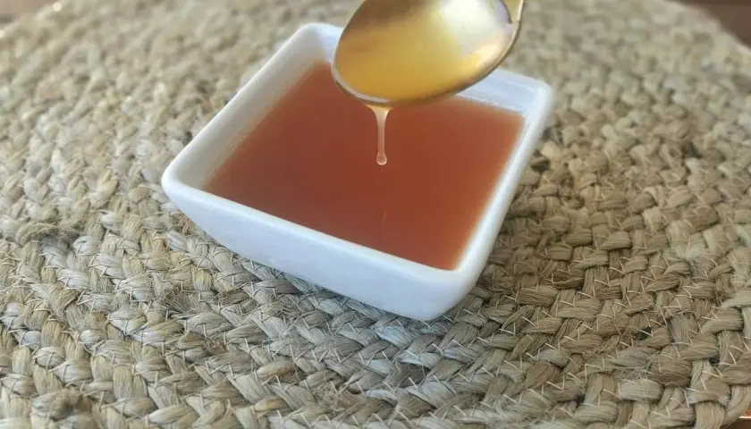 Una gota de salsa agridulce cae de una cuchara dorada a un cuenco blanco sobre una estera tejida.