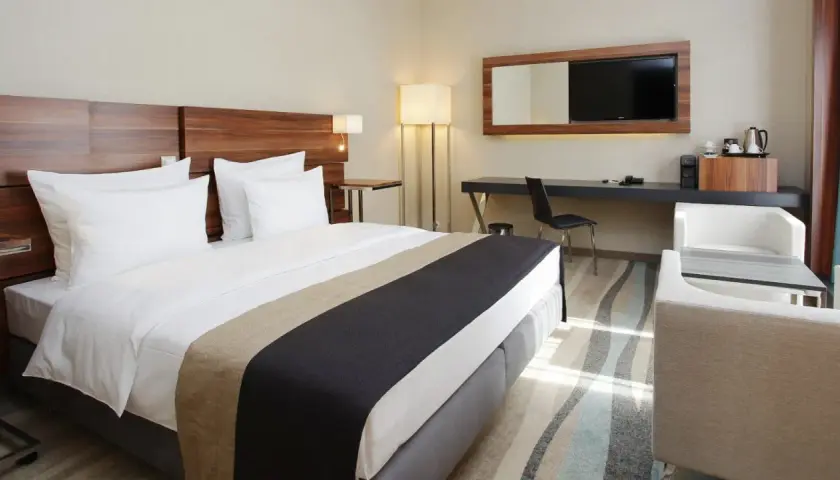 Nowoczesne wyposażenie pokoi hotelowych: wygodne łóżko, biurko z TV, fotele i stolik.