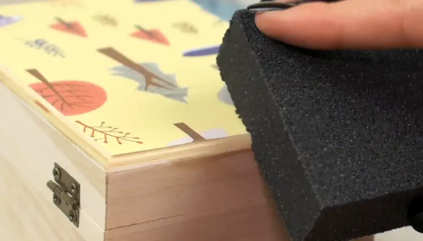 Aplicando pegamento para saber como pegar papel en madera, con una esponja sobre una caja decorada con hojas.