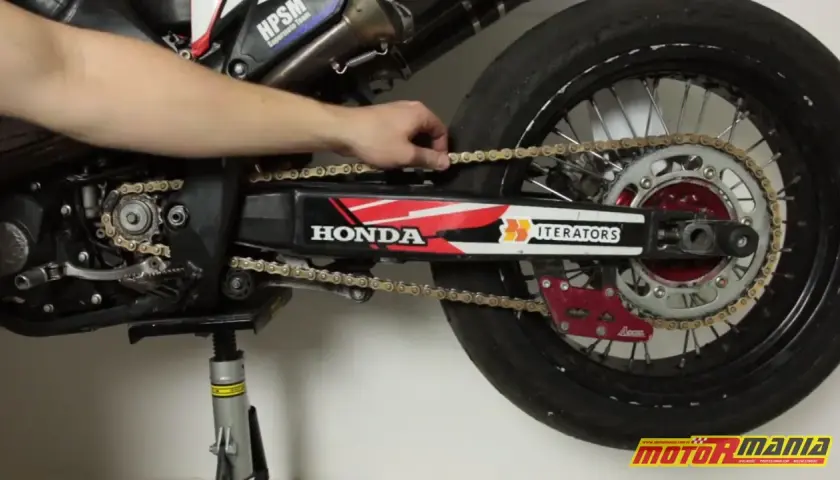 Ręka reguluje naciąg łańcucha w motocyklu Honda. Widać zębatkę, wahacz i tylne koło.