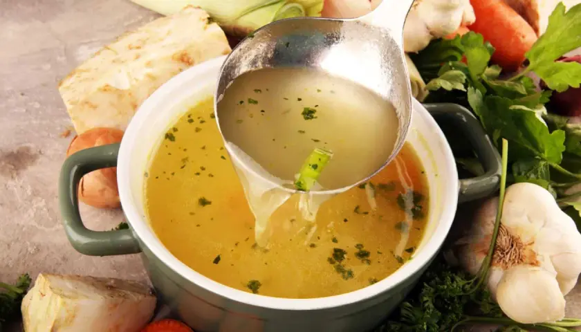 Un cucharón sirve un caldo depurativo, rodeado de verduras frescas como apio, zanahoria y ajo.