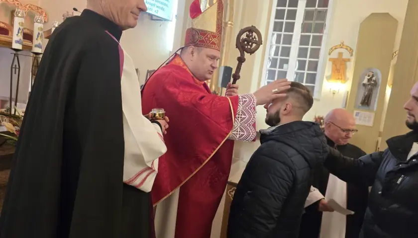 Biskup w czerwonej szacie udziela sakramentu bierzmowania młodemu mężczyźnie w kościele.