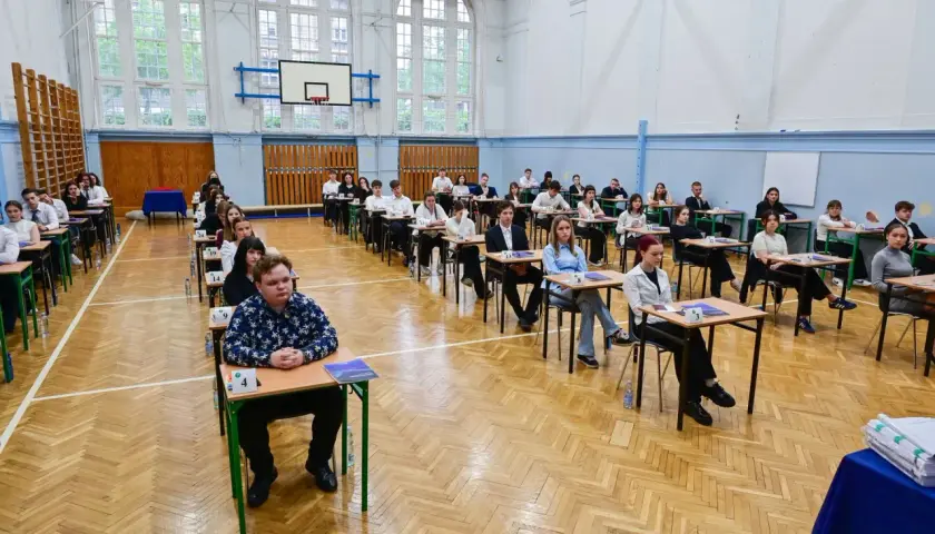 Uczniowie siedzą w ławkach w sali gimnastycznej, zdając egzamin. To typowy widok, gdy młodzi ludzie w wieku ok. 18 lat zdają maturę.