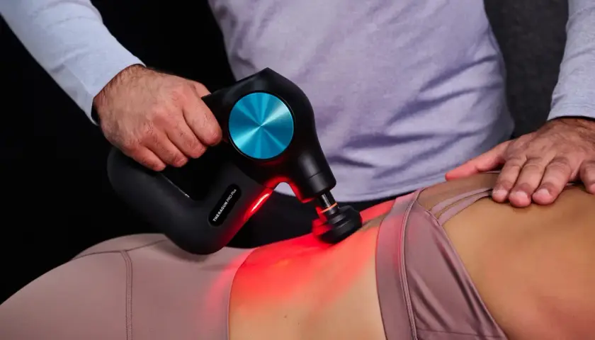 Eine Hand führt eine Massagepistole zur Anwendung am unteren Rücken einer Person. Rotes Licht leuchtet auf.