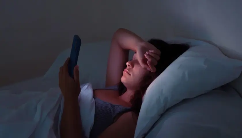 Kobieta leży w łóżku, przeglądając telefon w środku nocy. Jej ręka zasłania oczy, oznaka bezsenności.