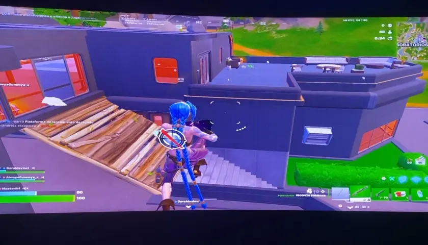 Postać z niebieskimi warkoczami w grze Fortnite buduje drewnianą rampę, celując w budynek. Opis gry Fortnite.