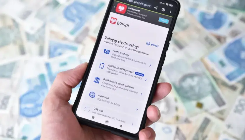 Dłoń trzyma smartfon z ekranem logowania do serwisu gov.pl. Dostępne opcje to m.in. Profil zaufany przez bankowość elektroniczną.