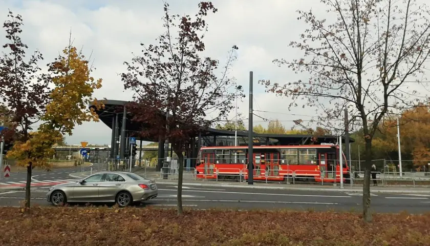 Srebrny samochód i czerwony tramwaj na przystanku, otoczone jesiennymi drzewami. Non stop big single.