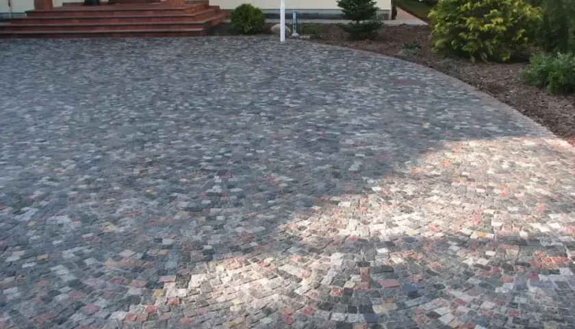 Piękny podjazd z kamienia, ułożony w łuk, prowadzi do drzwi domu. Wokół rosną drzewka i krzewy.