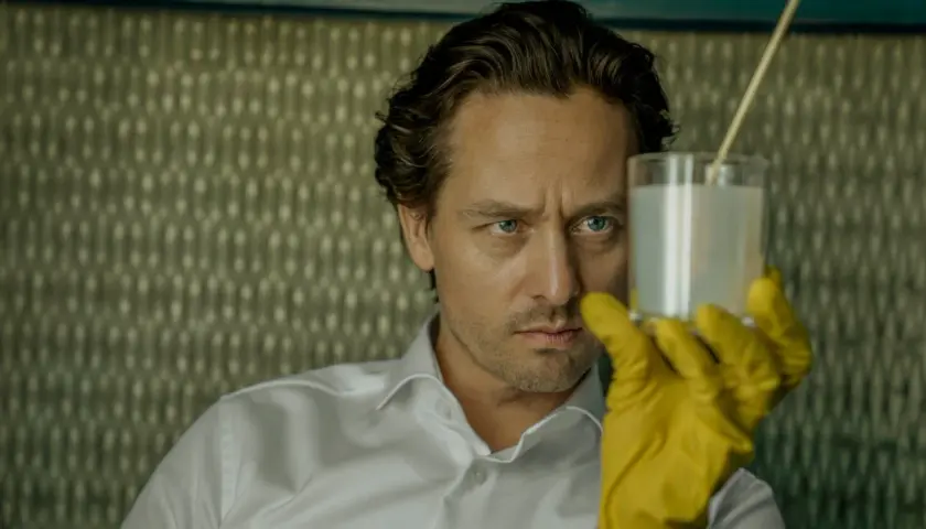 Ein Mann mit blauen Augen und gelben Handschuhen hält ein Glas mit weißer Flüssigkeit. Er ist Teil der "Achtsam Morden Reihe".
