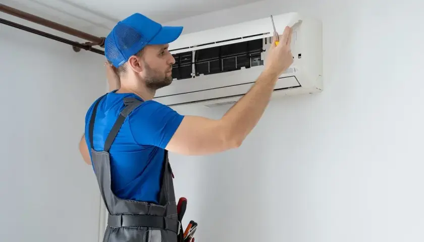 Technik w niebieskiej czapce i ogrodniczkach naprawia klimatyzator.