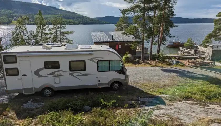 Un camping-car blanc est garé près d'un lac bordé de pins. Une petite cabane et un bateau sont visibles sur l'eau.