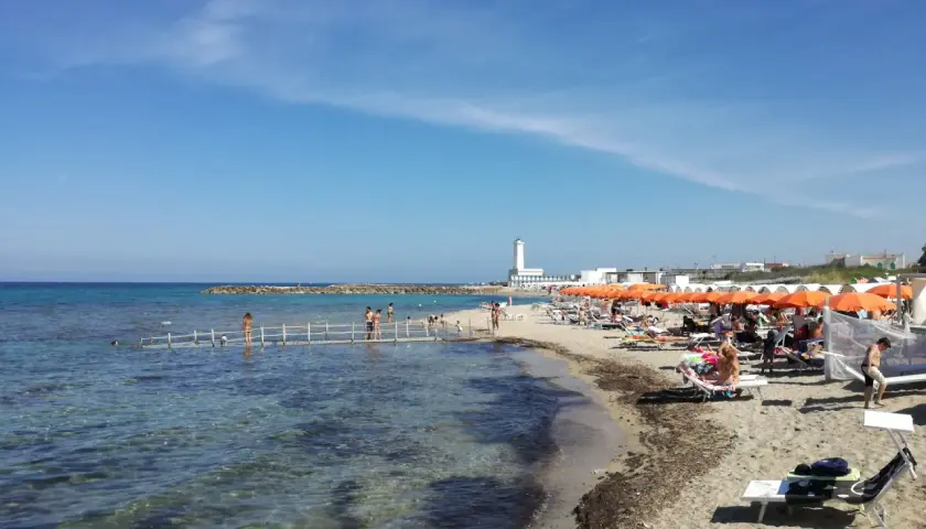 Plage animée à San Cataldo, Pouilles, avec des parasols orange, des baigneurs sur un ponton et un phare blanc en arrière-plan.