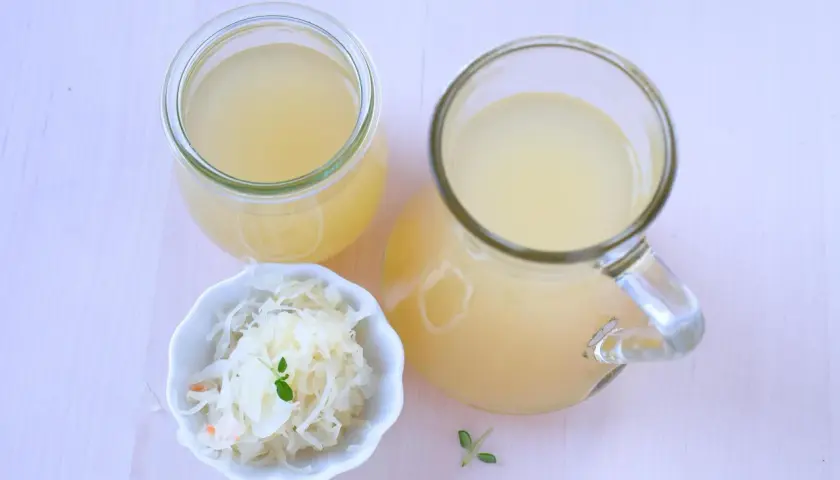 Ein Glas und ein Krug mit Sauerkrautsaft, reich an Vitaminen, stehen neben einer Schale Sauerkraut.