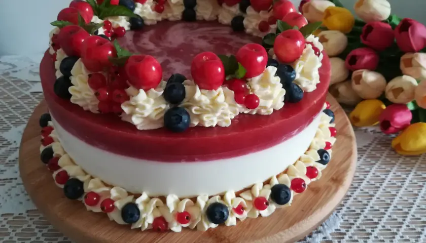 Wykwintny tort z warstwą białego kremu i błyszczącą, czerwona żelką truskawkową, ozdobiony bitą śmietaną, owocami i listkami mięty.