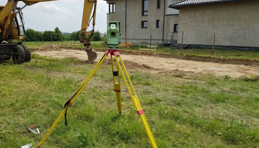 Geodeta z niwelatorem na statywie na placu budowy obok koparki i domów.