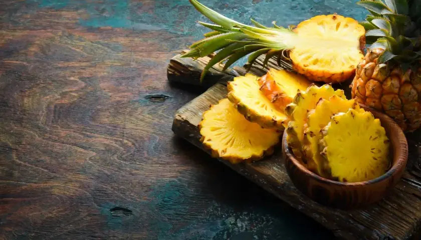 Frische Ananas-Scheiben, eine halbe Ananas und eine ganze Frucht liegen auf einem rustikalen Holzbrett. Das Fruchtfleisch ist leuchtend gelb und zeigt die typische Struktur, die auch für Bromelain bekannt ist.
