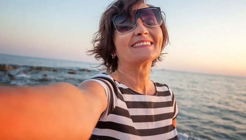Uśmiechnięta kobieta w okularach przeciwsłonecznych robi selfie na plaży o zachodzie słońca.