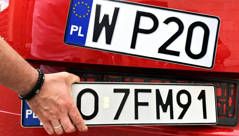 Ręka z bransoletką montuje tablicę rejestracyjną z napisem "O 7FM91" na czerwonym samochodzie. Powyżej widać inną tablicę "W P20".