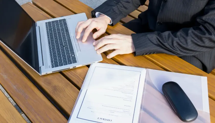 Biznesmen przy laptopie analizuje, ile kosztuje założenie księgi wieczystej. Na stole dokumenty i etui na okulary.