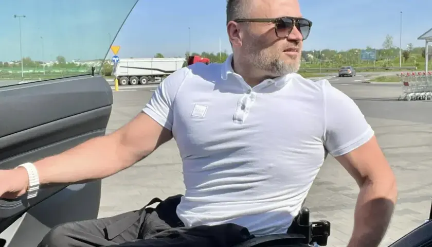 Mężczyzna na wózku inwalidzkim przy otwartych drzwiach samochodu na parkingu.