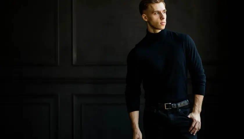 Młody mężczyzna w czarnym golfie i jeansach, pozujący na tle ciemnej ściany.