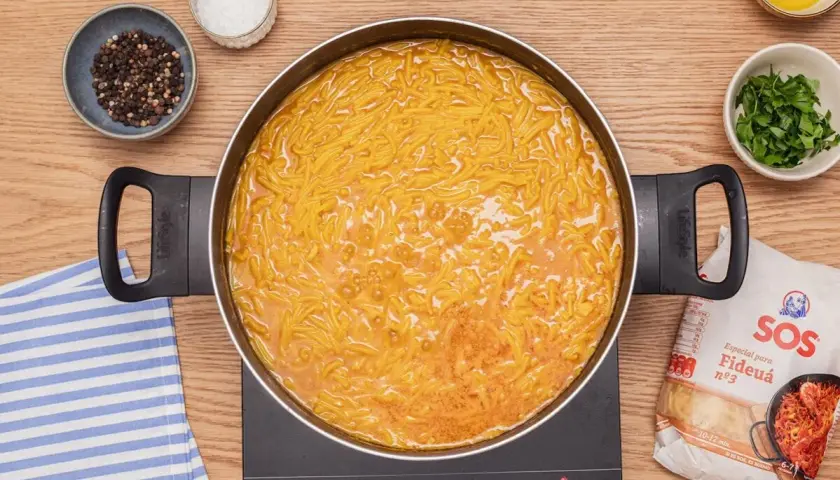 Fideuá con almejas burbujeando en una sartén. Alrededor, especias, perejil y un paquete de fideos.