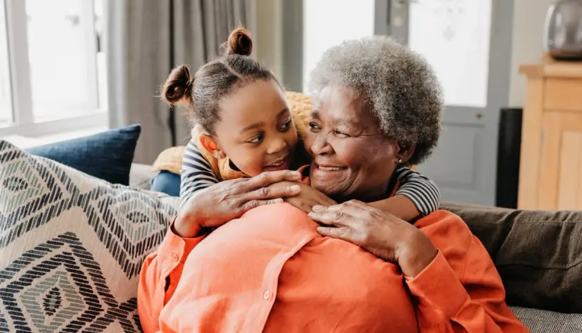 Ein süßer Moment zwischen Oma und Enkel. Das kleine Mädchen umarmt ihre Großmutter von hinten, beide lächeln sich liebevoll an.