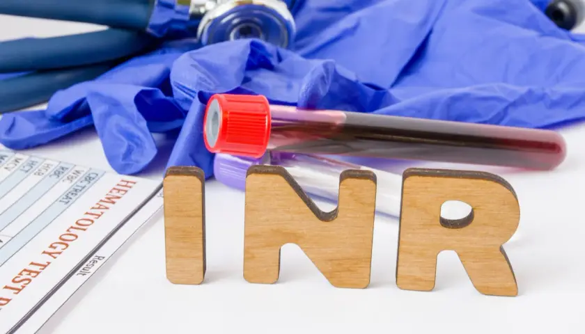 Drewniane litery INR na tle rękawiczek medycznych, stetoskopu i probówek z krwią. Wyniki badań hematologicznych.