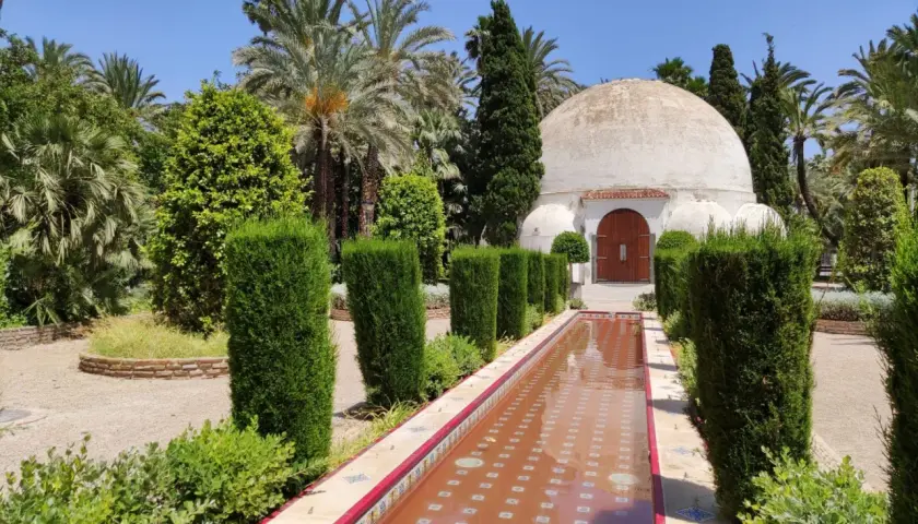 Un estanque de agua rojiza y un edificio abovedado en el parque el palmeral, rodeado de palmeras y vegetación exuberante.