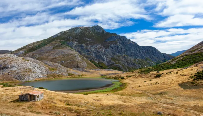 Pequeña cabaña de piedra junto a un lago ausente, rodeada de montañas escarpadas y prados secos bajo un cielo azul con nubes.
